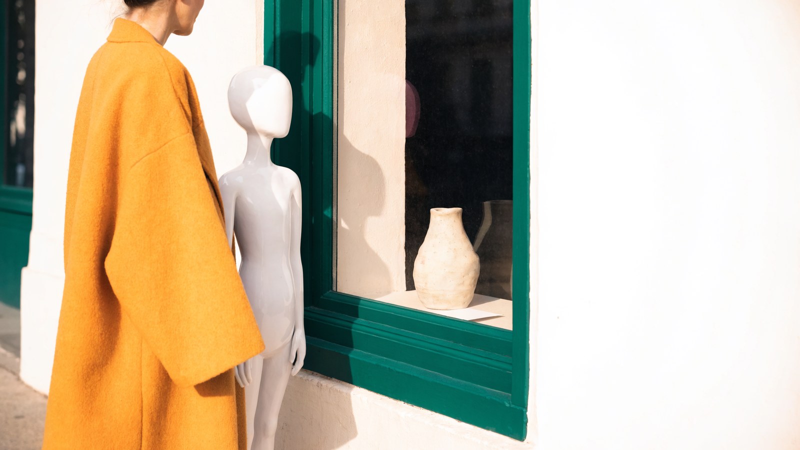 A woman in a mustard coat and a smooth, featureless white figure standing side by side on the pavement, both looking into a cream and deep-green shop window at a single ceramic vessel